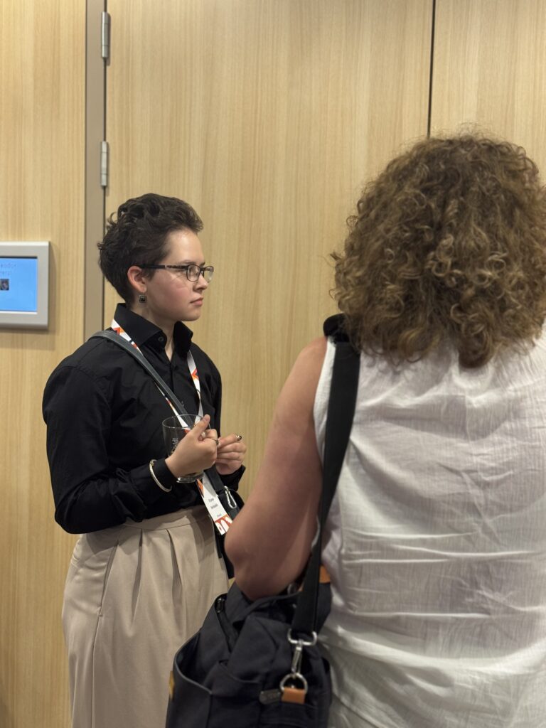 Woman with short dark hair and glasses wearing a black shirt and beige pants holds a glass and talks to another person with curly hair in a wood paneled room