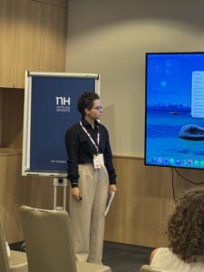 Young presenter in a black shirt stands beside a blue NH Hotels banner at a conference with a large screen showing a computer desktop to the right