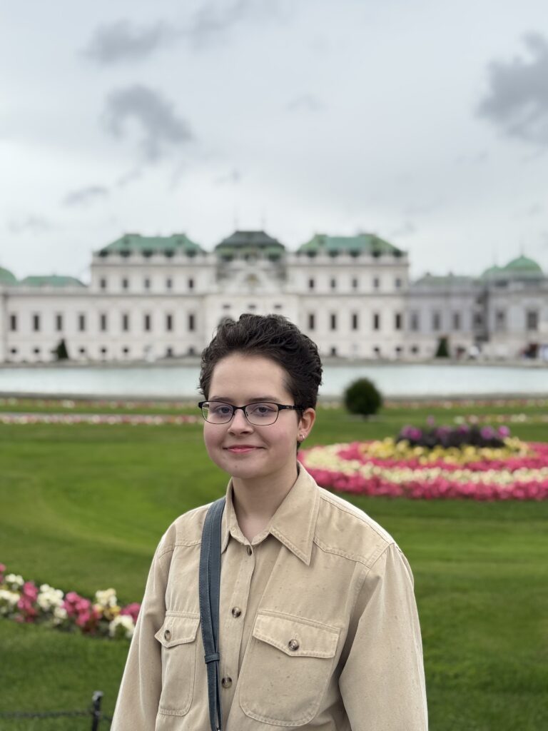Person with short dark hair and glasses wearing a beige shirt stands in a formal garden with a grand building blurred in the background