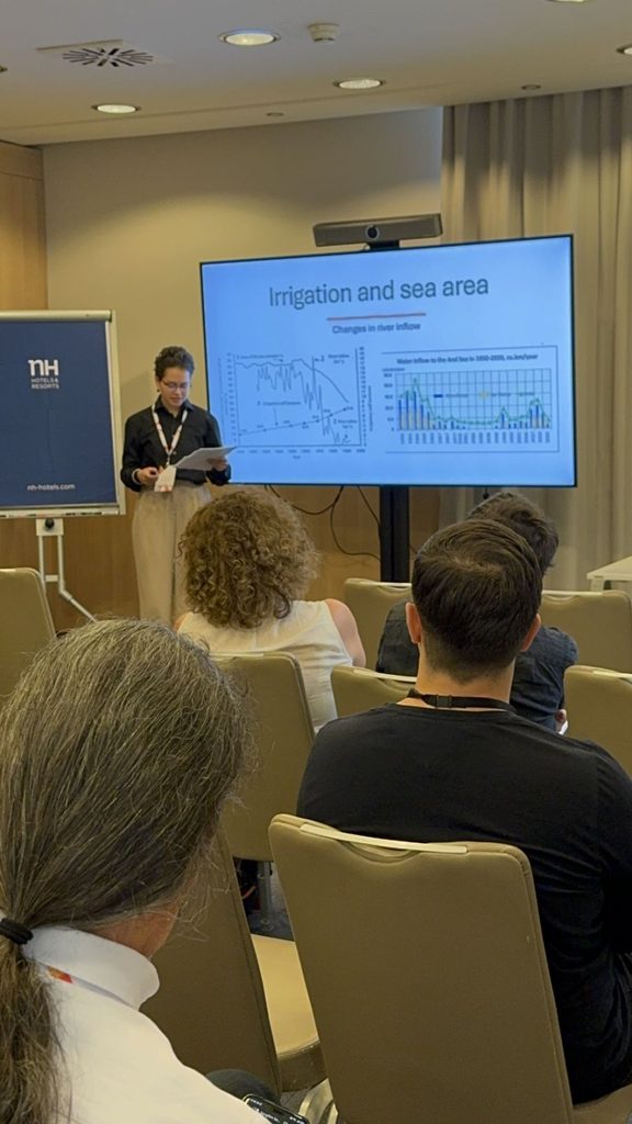Presenter standing beside a large screen displaying Irrigation and sea area with graphs audience seated in a conference room